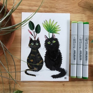 以下が含まれることがあります： 緑色の目をした黒い猫2匹の水彩画です。1匹の猫の頭には緑の葉の植物が生えていて、もう1匹の猫の頭にも緑の葉の植物が生えています。猫は白い背景に座っています。