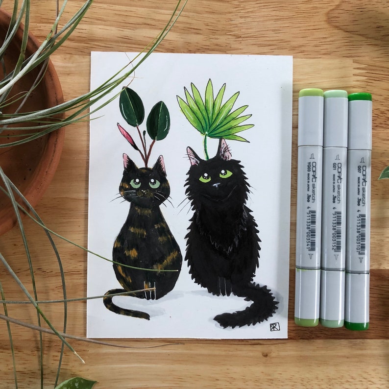 以下が含まれることがあります： 緑色の目をした黒い猫2匹の水彩画です。1匹の猫の頭には緑の葉の植物が生えていて、もう1匹の猫の頭にも緑の葉の植物が生えています。猫は白い背景に座っています。