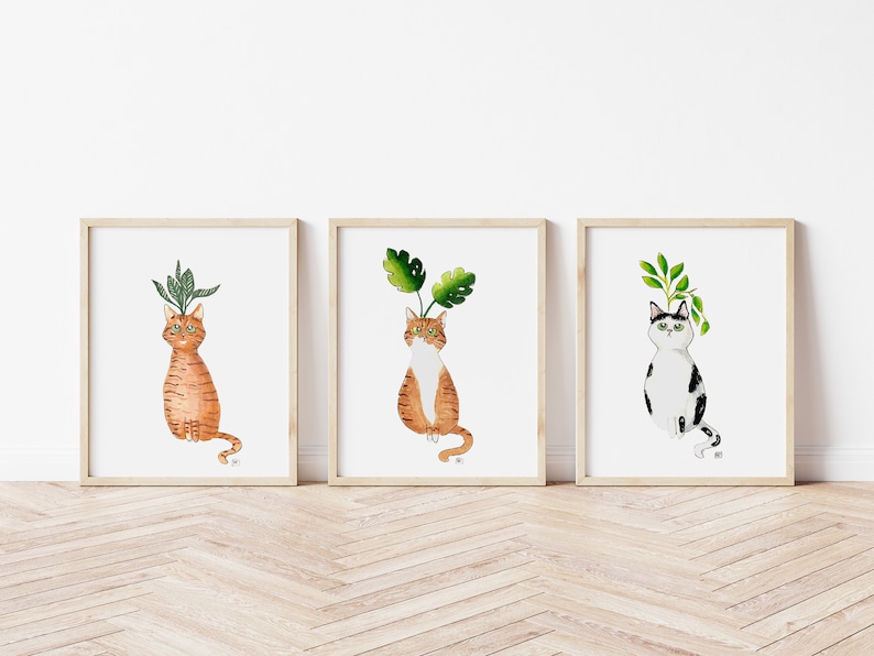 以下が含まれることがあります： 頭から植物が生えている猫の水彩画のイラストが3つ額装されています。猫は座って鑑賞者を見ています。最初の猫はオレンジと白、2番目の猫はオレンジと白、3番目の猫は黒と白です。