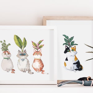 以下が含まれることがあります： 頭から植物が生えている猫を描いた水彩画が3枚、額装されています。猫は直立して座り、鑑賞者を見つめています。絵画は白い額縁に入っていて、白い棚に飾られています。