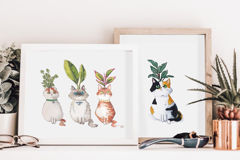 以下が含まれることがあります： 頭から植物が生えている猫を描いた水彩画が3枚、額装されています。猫は直立して座り、鑑賞者を見つめています。絵画は白い額縁に入っていて、白い棚に飾られています。