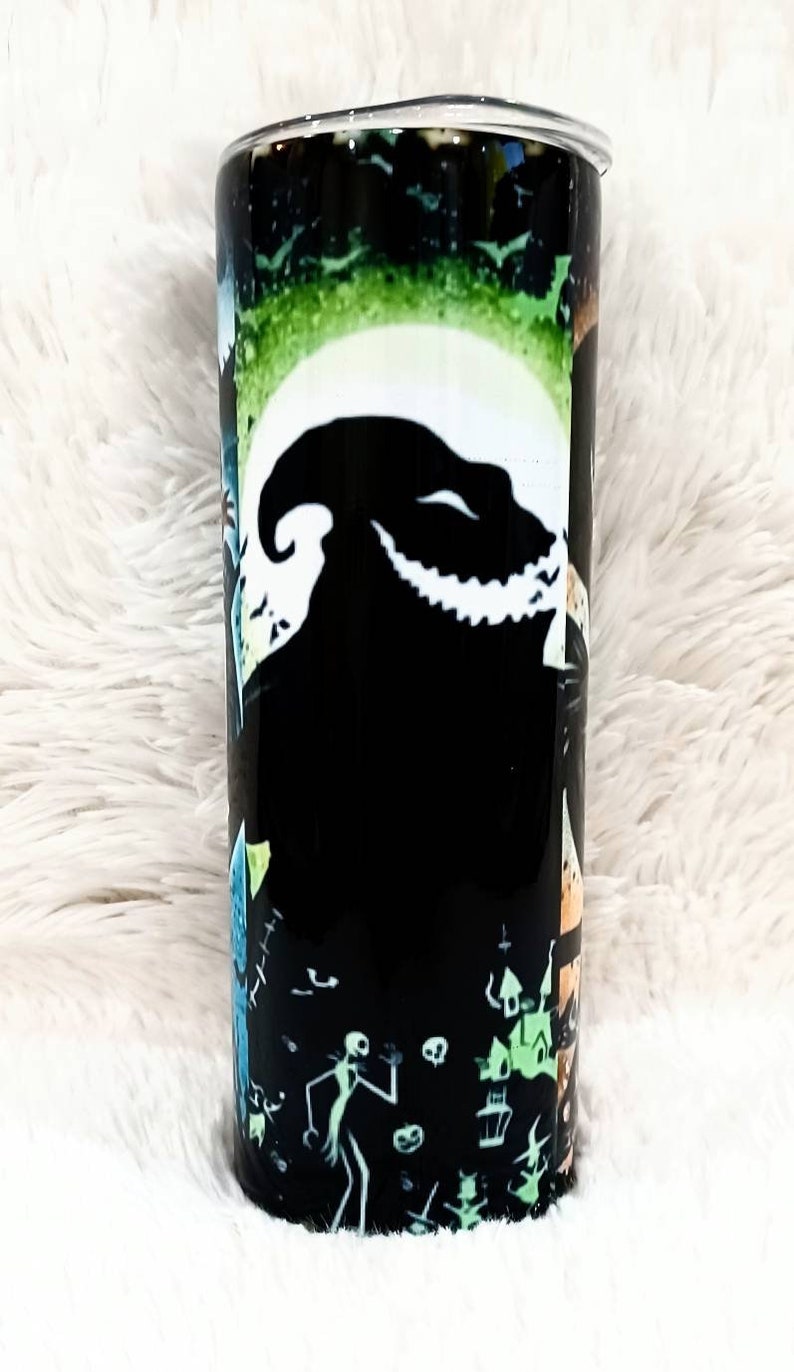 Puede incluir: Un vaso cil&iacute;ndrico con un dise&ntilde;o de Pesadilla antes de Navidad. El vaso negro tiene una luna blanca, una silueta de Oogie Boogie e im&aacute;genes de Jack Skellington y otros personajes. El fondo tiene un degradado verde y negro.