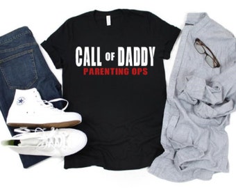 May include: A black t-shirt with the text "CALL OF DADDY PARENTING OPS" in white and red. The shirt is paired with blue jeans, white sneakers, and a gray button-down shirt with glasses.