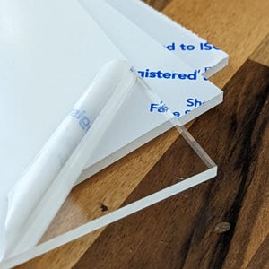 Op de afbeelding: Close-up van heldere acrylplaten met gedeeltelijk verwijderde beschermfolie. De platen zijn op elkaar gestapeld en tonen een transparante rand. De beschermfolie heeft blauwe tekst met "registered" en "d to IS".