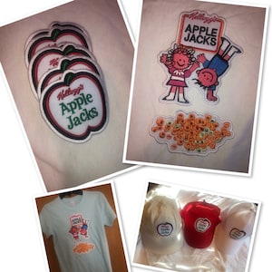 May include: A set of four iron-on patches featuring the Kellogg's Apple Jacks logo. The patches are white with red, green, and black embroidery. The patches are arranged in a stack, with the largest patch on top. The patches are on a white background.