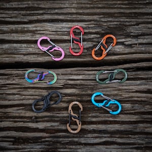 Puede incluir: Surtido de mosquetones S-Biner coloridos en varios colores, incluyendo rosa, rojo, naranja, iridiscente, verde, negro, beige y azul. Cada mosquetón presenta un diseño de doble puerta con una barra central. Los mosquetones están sobre una superficie de madera envejecida.