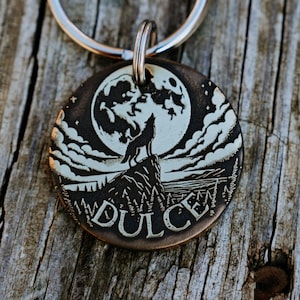May include: A round, bronze dog tag featuring a wolf howling at the moon. The tag has a silver ring attached and the word "Dulce" engraved on the bottom.