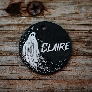 Puede incluir: Chapa redonda negra para perro con una ilustración de fantasma blanca y el nombre "CLAIRE" escrito en blanco.