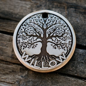 Op de afbeelding: Zilverkleurig metalen label met een levensboomontwerp en het woord "WILLOW" gegraveerd aan de onderkant.