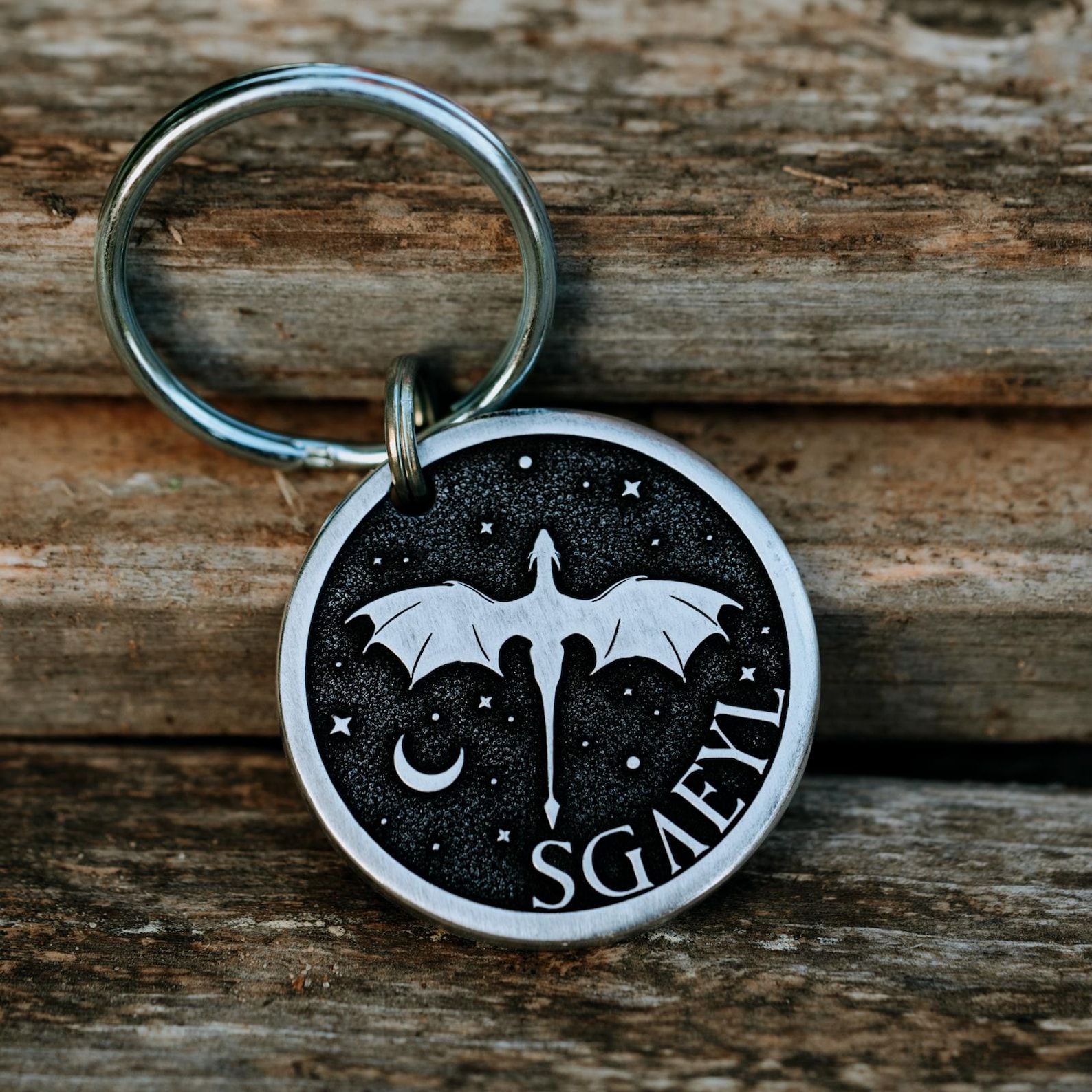 Round metal pet tag with dragon silhouette, stars, and a crescent moon on a black background. Text reads "SGAEYL."