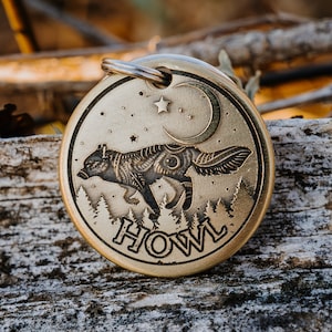 Puede incluir: Una chapa de perro de latón con un diseño de lobo, una luna creciente y estrellas. La chapa dice "HOWL" en una fuente estilizada.