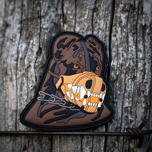 May include: A brown and black camouflage dog patch with a yellow and orange muzzle and a silver chain. The patch is designed to look like a dog wearing a muzzle.