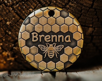 Puede incluir: Una placa circular de latón para mascotas con un patrón de panal en relieve en la superficie. El centro de la placa está grabado con el nombre Brenna en una fuente clara, situado sobre una ilustración detallada de una abeja melífera.