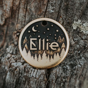 Puede incluir: Una chapa de perro de latón con un fondo oscuro y una silueta de bosque. El nombre "Ellie" está grabado en una fuente en negrita con una luna creciente y estrellas.