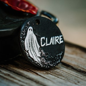 May include: Black round tag with a white ghost illustration and the name "CLAIRE" written in white lettering.