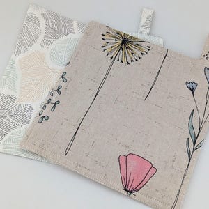 Könnte beinhalten: Zwei quadratische Topflappen aus Stoff. Einer hat einen beigen Hintergrund mit einem Löwenzahn- und Blumendesign in Schwarz, Gelb, Rosa und Blau. Der andere hat einen weißen Hintergrund mit einem Blattmuster in Grau, Beige und Hellblau.