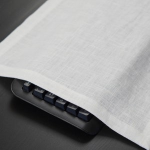 Könnte beinhalten: Ein weißes rechteckiges Platzset aus Leinen mit feiner Textur, das teilweise eine schwarze Tastatur bedeckt. Das Platzset hat ein schlichtes, minimalistisches Design mit einem leicht erhöhten Rand. Die Tastaturtasten sind schwarz.