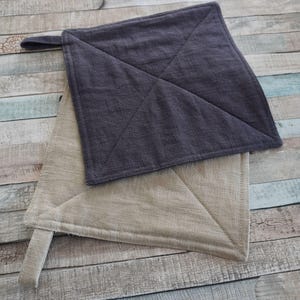 Könnte beinhalten: Zwei gepolsterte Topflappen, einer in hellbeige und einer in dunkelgrau. Beide Topflappen sind aus einem strukturierten Stoff gefertigt und haben eine quadratische Form.