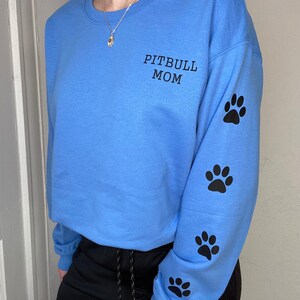 Può includere: Felpa azzurra con la scritta "PITBULL MOM" in nero sul petto sinistro. La manica destra presenta quattro impronte di zampe nere. Uno stile casual e confortevole.