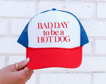 May include: A white, blue, and red trucker hat with the text "BAD DAY to be a HOT DOG" in red.