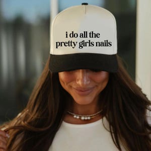 May include: A white and black trucker hat with the text "i do all the pretty girls nails" in black. The hat has a black brim and a white front panel. The hat is worn by a person with long brown hair.
