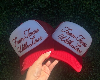 May include: Two red trucker hats with white front panels. The hats have the text "From Texas With Love" embroidered in a cursive font. The hats are held up against a green background.