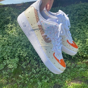 Puede incluir: Zapatillas blancas de ca&ntilde;a baja con un dise&ntilde;o de cono de helado. Las zapatillas tienen un cono de waffle marr&oacute;n, helado naranja y chispas de colores.