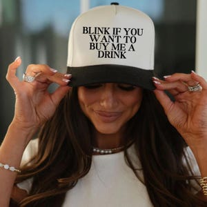 May include: A white and black baseball cap with the text "Blink if you want to buy me a drink" in black lettering.