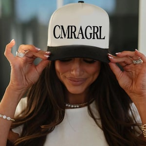 May include: A white and black baseball cap with the text "CMRAGRL" in black block letters. The cap has a black brim and a white crown. The person in the image is wearing the cap.