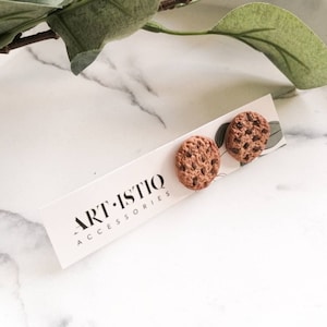Puede incluir: Un par de pendientes de botón marrones con forma de galleta y detalles de chispas de chocolate. Los pendientes se muestran en una tarjeta blanca con el texto "ART-ISTIQ ACCESSORIES". El fondo presenta una superficie veteada y hojas verdes.