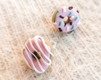 May include: Two miniature donut earrings. One donut has pink frosting with white icing stripes. The other donut has pink frosting with green and black sprinkles. The earrings are on a textured, off-white surface.