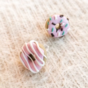 Puede incluir: Dos pendientes en miniatura con forma de donut. Uno tiene glaseado rosa con rayas de glaseado blanco. El otro tiene glaseado rosa con chispas verdes y negras. Los pendientes están sobre una superficie texturizada blanquecina.