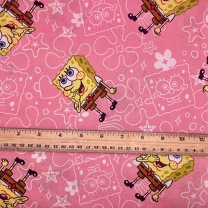 May include: Pink fabric with a repeating pattern of SpongeBob SquarePants, and white outlines of stars, hearts, and other shapes. A wooden ruler is placed across the fabric, measuring up to 28 cm.