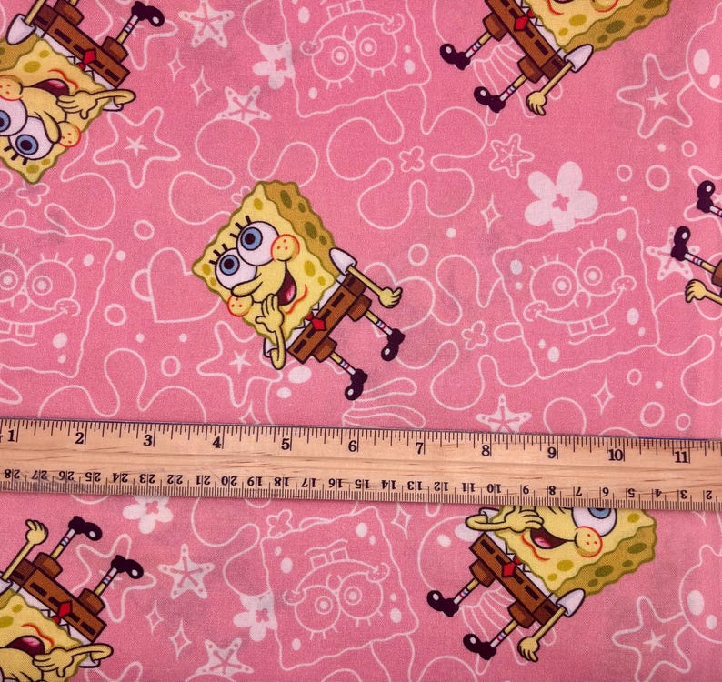 May include: Pink fabric with a repeating pattern of SpongeBob SquarePants, and white outlines of stars, hearts, and other shapes. A wooden ruler is placed across the fabric, measuring up to 28 cm.