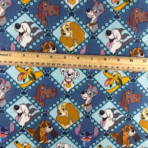 May include: Blue fabric featuring a repeating pattern of cartoon dog characters in diamond shapes. The dogs are in various colors and poses, with a ruler placed across the fabric. The ruler measures up to 11 inches.