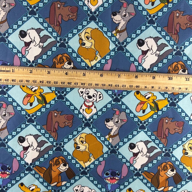 May include: Blue fabric featuring a repeating pattern of cartoon dog characters in diamond shapes. The dogs are in various colors and poses, with a ruler placed across the fabric. The ruler measures up to 11 inches.
