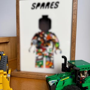 May include: A framed print with the word "SPARES" above a blurred Lego figure with a colourful pattern. A green and yellow Lego tractor and a yellow Lego construction vehicle are in the foreground.