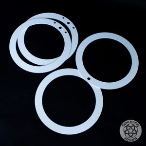 Pode incluir: Vários objetos circulares brancos com um ou mais furos são dispostos sobre um fundo preto. Os objetos parecem ser feitos de um material sólido e têm uma superfície lisa. Os objetos são de tamanhos variados e sobrepõem-se.