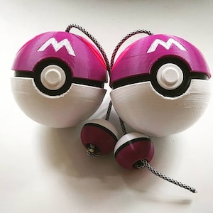 May include: Two pink and white Poké Balls with a black 'M' on the top half of each ball. The balls are connected by a black and white string.