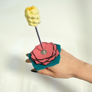 May include: A unique ring featuring a layered pink and teal flower design. A yellow, textured element is attached to the top of the ring with a thin, purple rod. The ring is held by a hand against a white background.