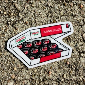 May include: A die-cut sticker featuring a white cardboard box with red accents containing twelve black and red circular doughnut illustrations. The box lid displays the text Krispy Kreme and Original Glazed, with the text Lindsays Paperworks visible.