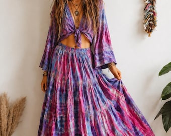 Puede incluir: Un conjunto de dos piezas compuesto por una blusa de manga larga con nudo frontal y una falda larga escalonada a juego. Ambas prendas presentan un vibrante estampado tie-dye en tonos magenta, morado y azul, creando un conjunto muy colorido.
