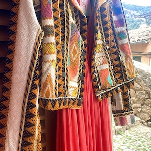 Puede incluir: Una chaqueta colorida estampada con botones negros cuelga junto a un vestido largo y fluido de color coral. La chaqueta presenta un diseño geométrico con detalles en naranja, amarillo y negro. Una etiqueta con la palabra "LUZ" es visible.