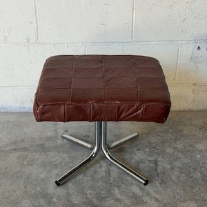 May include: A square ottoman featuring a dark brown leather top constructed from a grid of stitched square patches. The seat is supported by a four-pronged chrome metal base with black plastic caps at the end of each leg, providing a stable foundation.