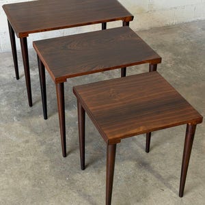 May include: A set of three nesting tables crafted from dark brown wood with a prominent grain pattern. Each table features a rectangular top and four tapered legs, arranged in a cascading formation from the largest to the smallest size.