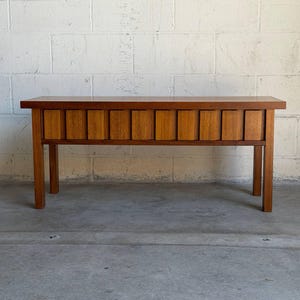 May include: A rectangular wooden bench with a warm brown finish, featuring a flat top surface and four straight legs. The front apron is composed of a series of vertical, slightly protruding wooden slats that create a textured, rhythmic pattern.