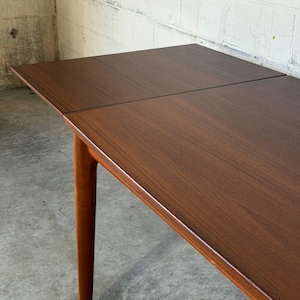 May include: A rectangular wooden dining table with a rich brown finish and visible wood grain. The table features a smooth, flat surface with a visible seam indicating an extendable leaf, supported by a sturdy, tapered cylindrical wooden leg.