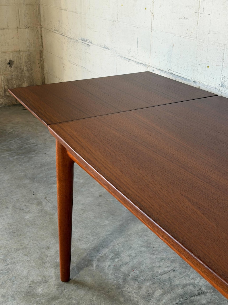 May include: A rectangular wooden dining table with a rich brown finish and visible wood grain. The table features a smooth, flat surface with a visible seam indicating an extendable leaf, supported by a sturdy, tapered cylindrical wooden leg.