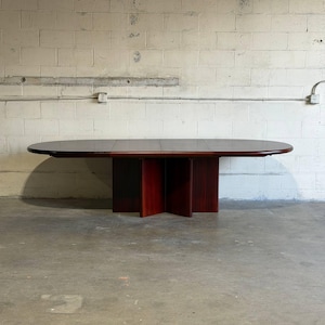 May include: A dark wood dining table with an elongated oval top and a central pedestal base featuring four intersecting vertical panels. The table surface has a smooth, polished finish with visible wood grain, supported by a sturdy, cross-shaped base.