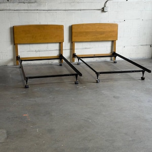 May include: Two matching twin bed frames featuring light brown wooden headboards with a curved top edge. Each frame is constructed from black metal rails and includes four small caster wheels at the base for mobility and support.